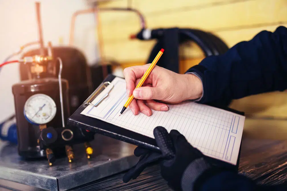 Technician's hands holding a clipboard and making notes in front of a heat pump system.